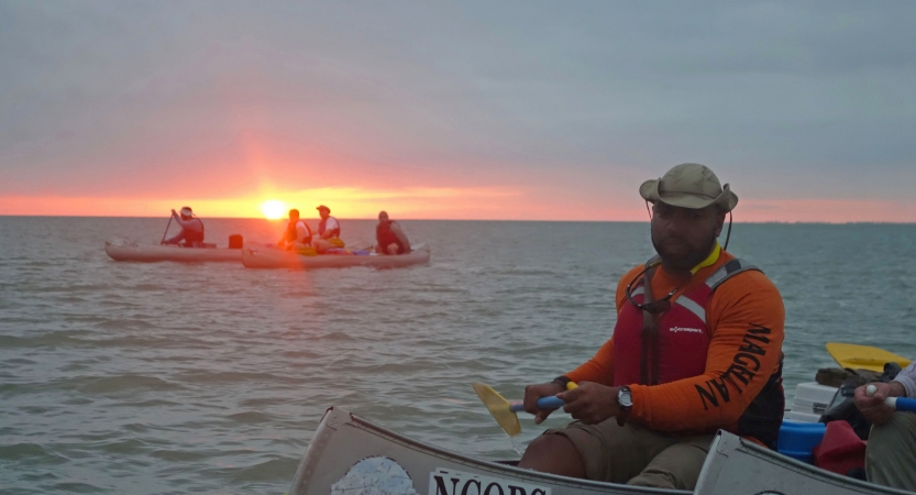 A group of people paddle canoes on open water as the sun sets on the horizon.